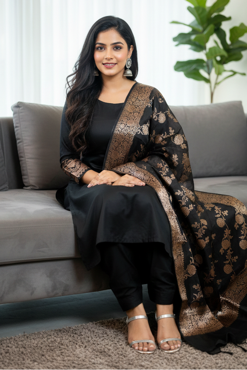 Woman sitting on a couch wearing a black and gold traditional outfit with a plant in the background.