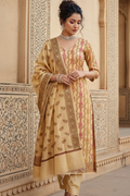 Woman in a traditional beige and gold embroidered outfit standing in front of ornate wall.