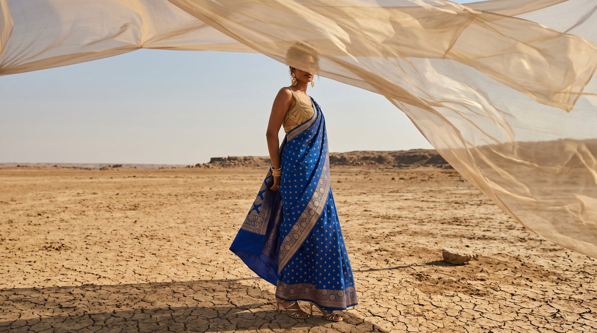 A woman stands in a dry desert landscape wearing a blue patterned sari with a beige blouse, under a large flowing sheer cream fabric drape.